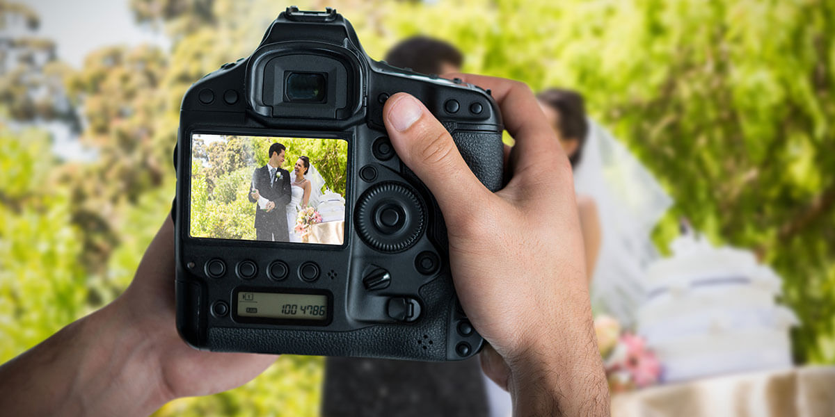 Cropped image of hands holding camera against newlywed couple celebrating with opening champagne bottle at park