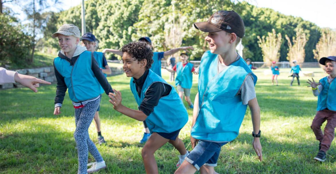 A group of four young children wearing blue vests race one another in a green park