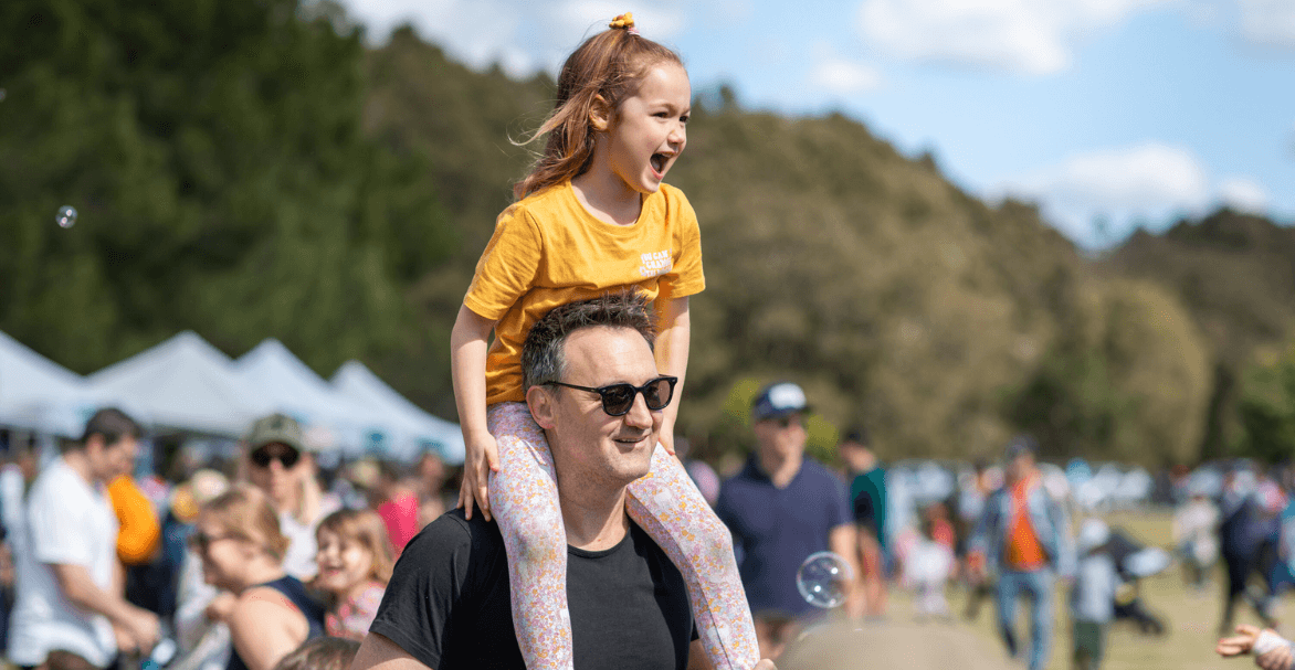 A young girl sits on a man's shoulders smiling at an outdoor event in Centennial Park