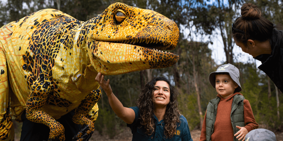 A life-sized yellow dinosaur meets a woman with a young boy wearing a hat and his mother