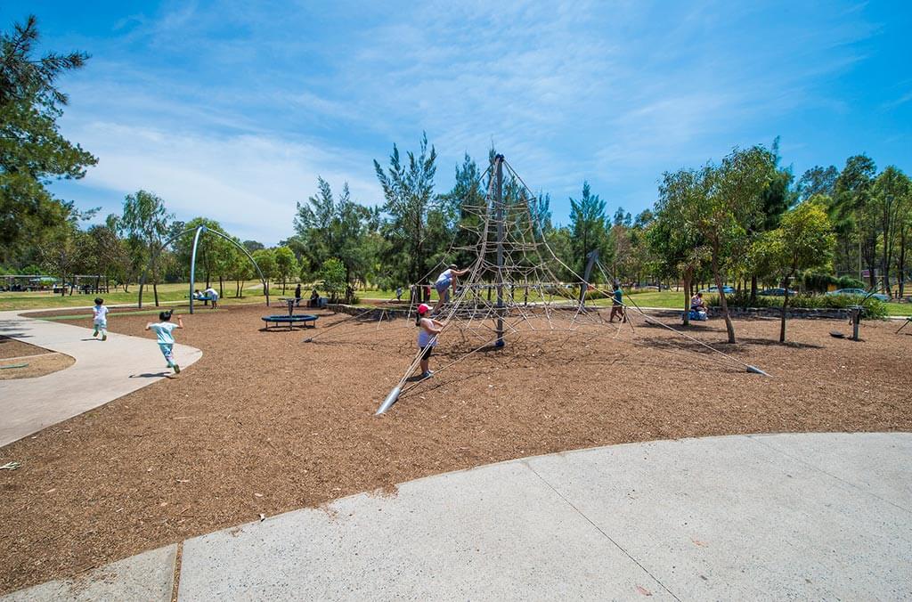 A vibrant park scene with a playground, swings swaying, and a tall tree offering shade for children.