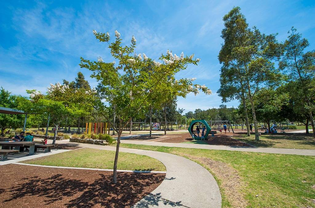A vibrant park scene with a colorful playground, lush green grass, and shady trees.