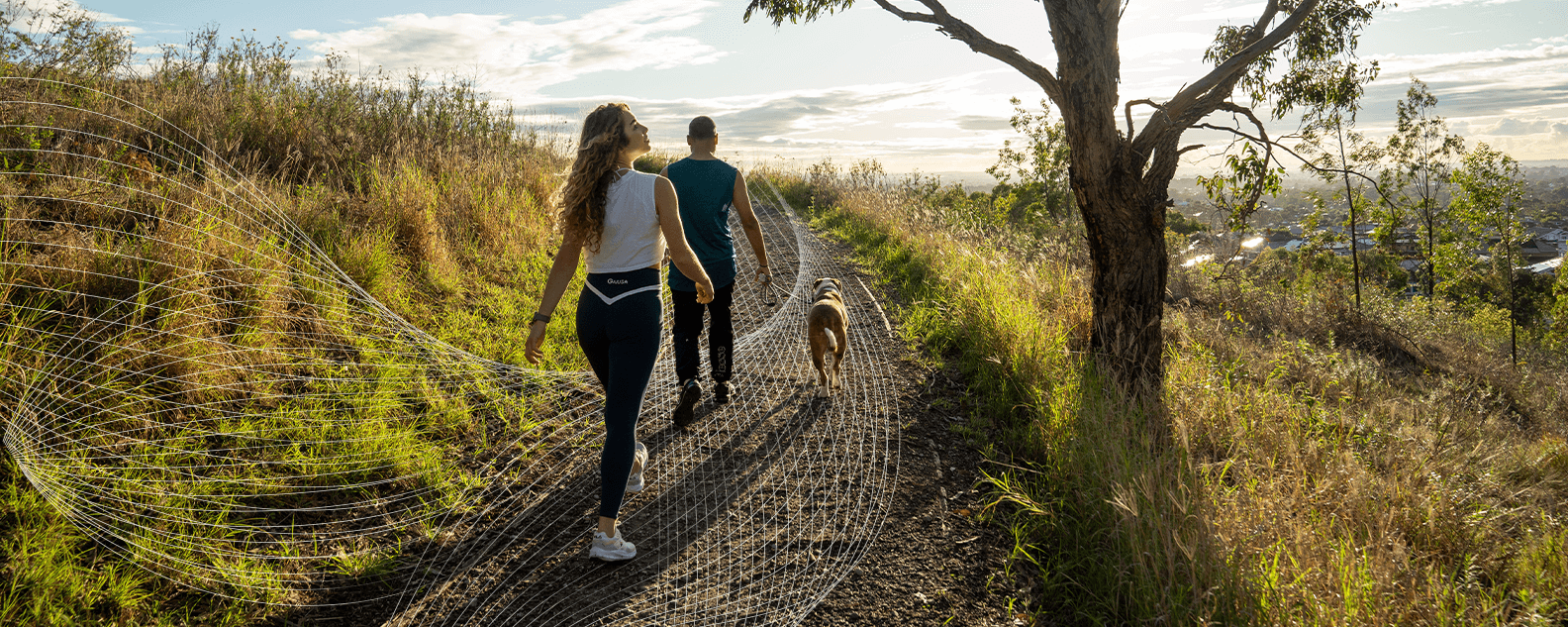 A couple and their dog walk on a dirt path through a grassy, sunlit landscape, with a tree on the right and a town visible in the distance.