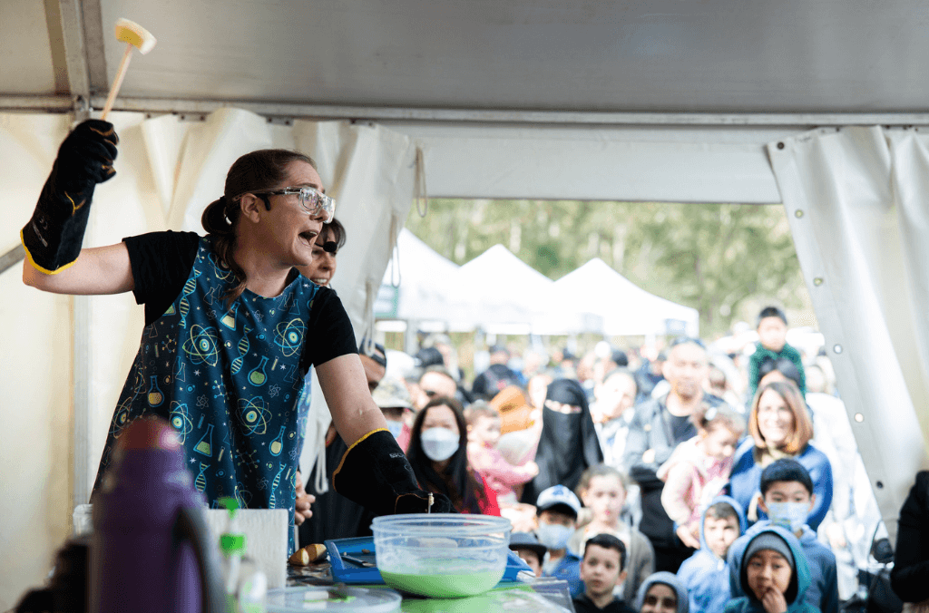 A female scientist wearing safety goggles performs a science experiment on an outdoor stage in front of a live crowd.