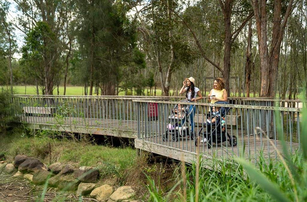 Two women with children in prams stand on a wooden bridge surrounded by trees in a peaceful forest setting.