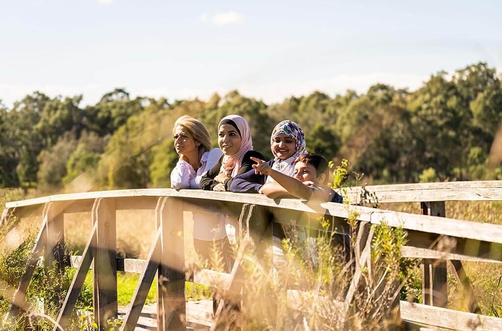 Three women are standing on a wooden bridge, chatting and laughing as they admire the nature surrounding them.
