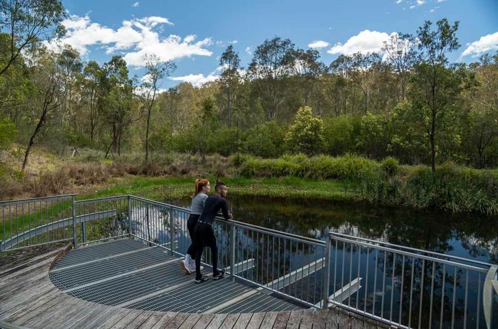 Two people stand on a wooden bridge, overlooking a serene pond at Plough and Harrow Loop, surrounded by nature.